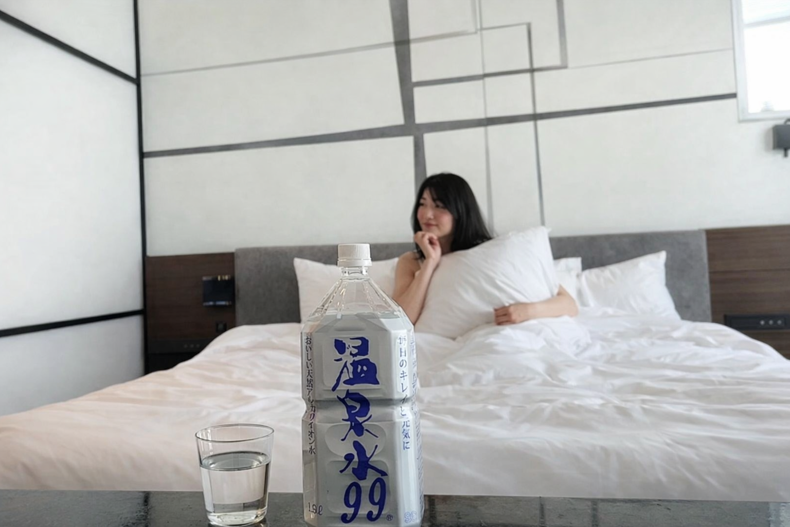 ホテルのベッドでくつろぐ女性とテーブルに置かれた温泉水99の1.9Lボトル