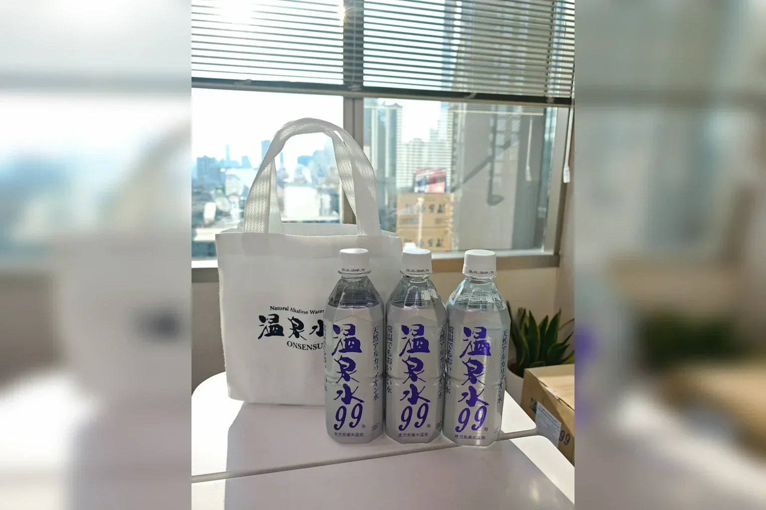 ONSENSUI99 sample water bottles and original tote bag displayed in the Thailand office with city view background