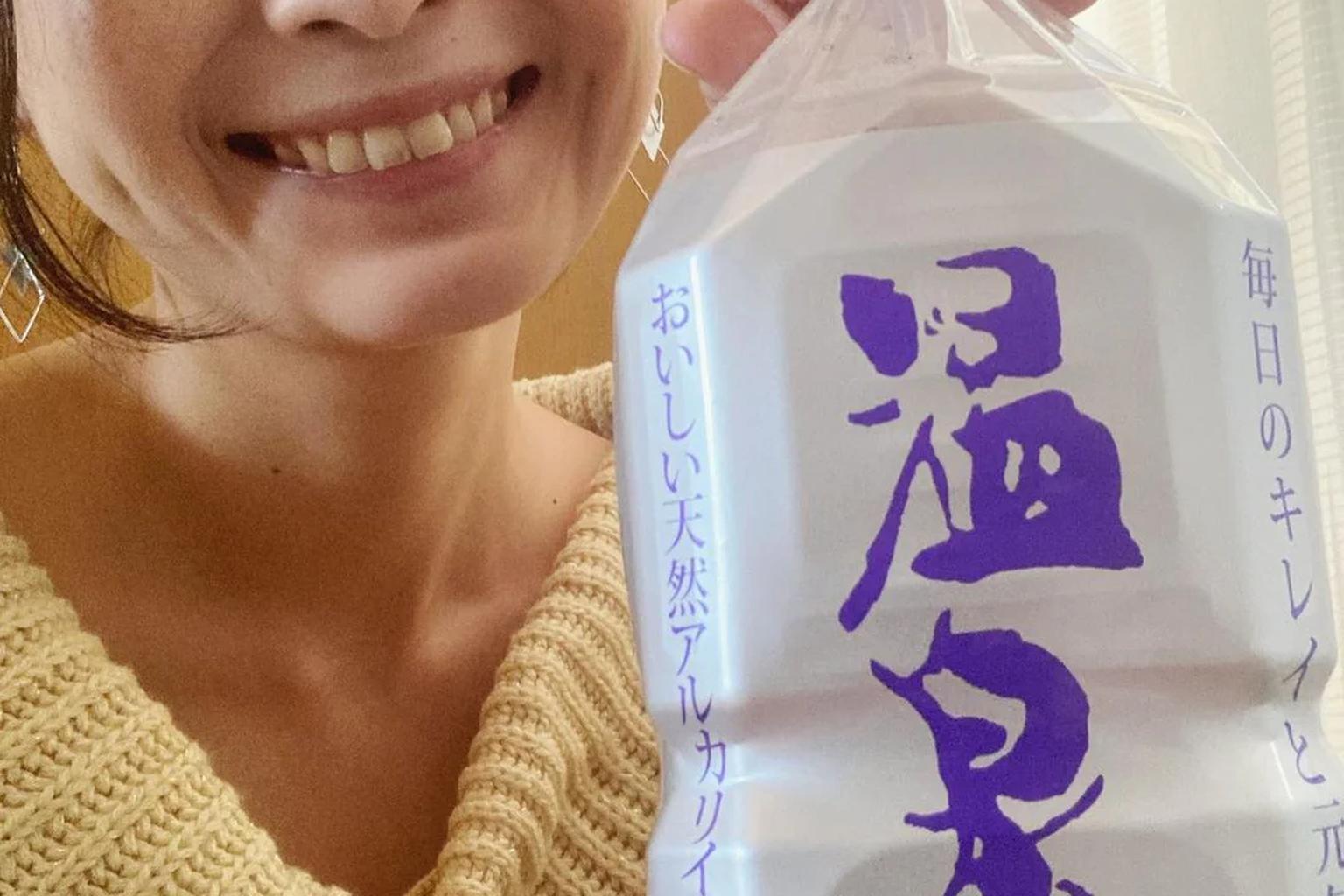 A smiling woman holding a bottle of ONSENSUI 99 natural alkaline water in warm daylight.