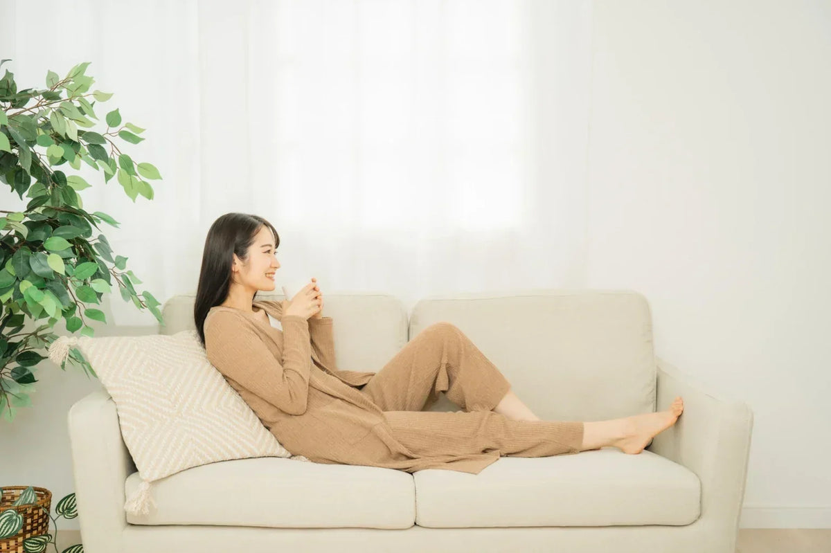 A woman relaxing on a sofa and drinking water, representing a comfortable daily hydration habit that supports healthy skin from within.
